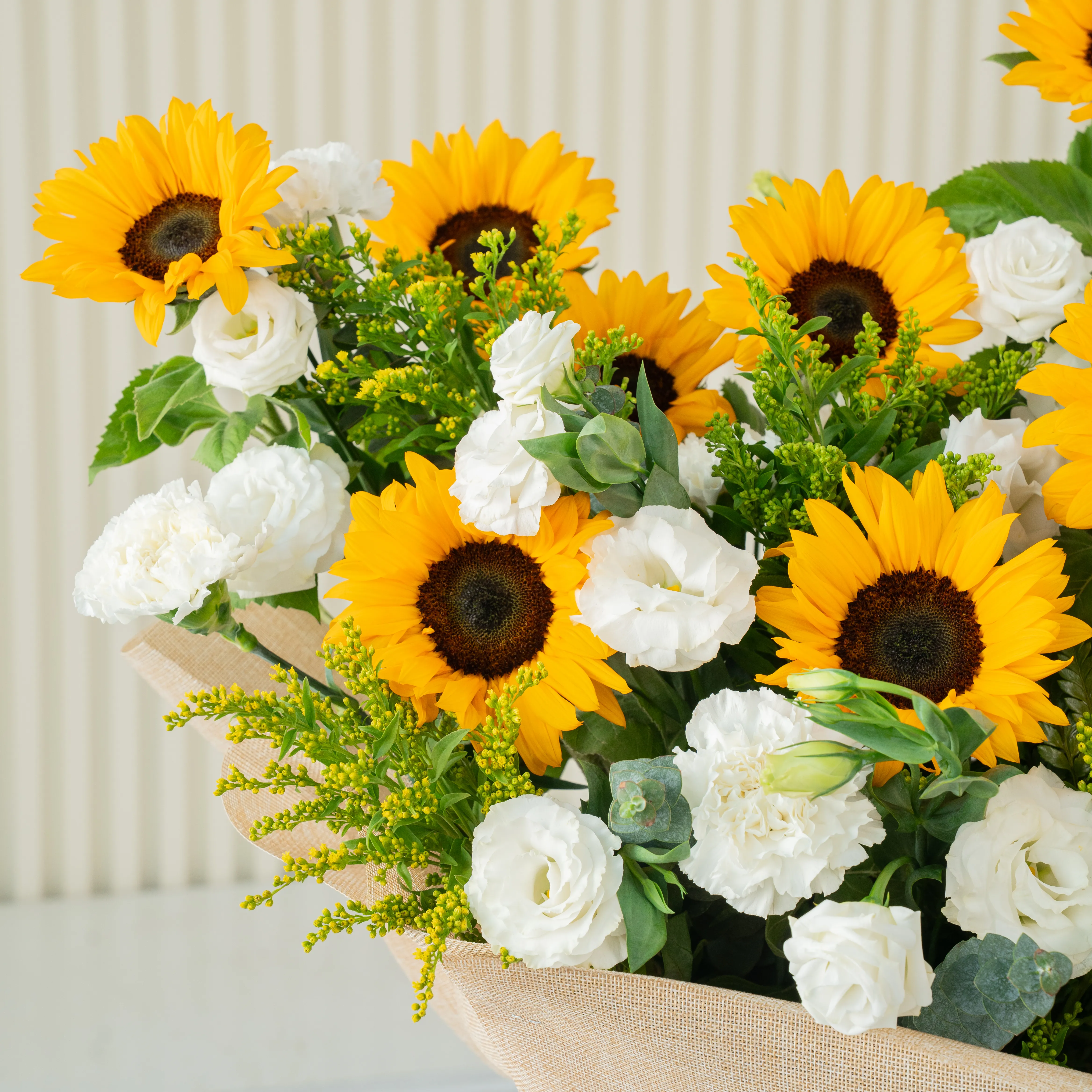 Bouquet of 11 sunflowers and 10 white lisianthus with green fillers and yellow statis in brown paper packing tied with white ribbon.