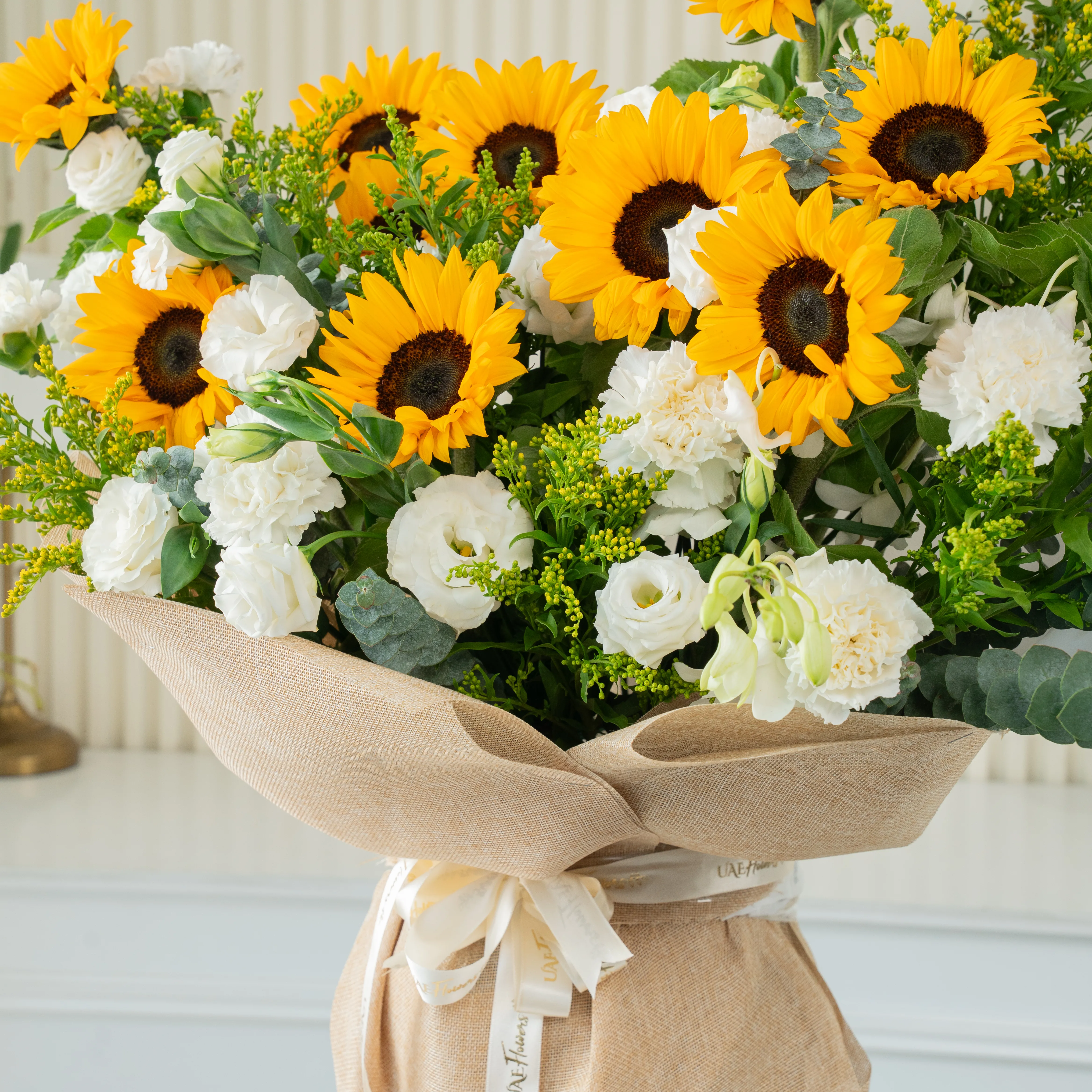 Bouquet of 11 sunflowers and 10 white lisianthus with green fillers and yellow statis in brown paper packing tied with white ribbon.
