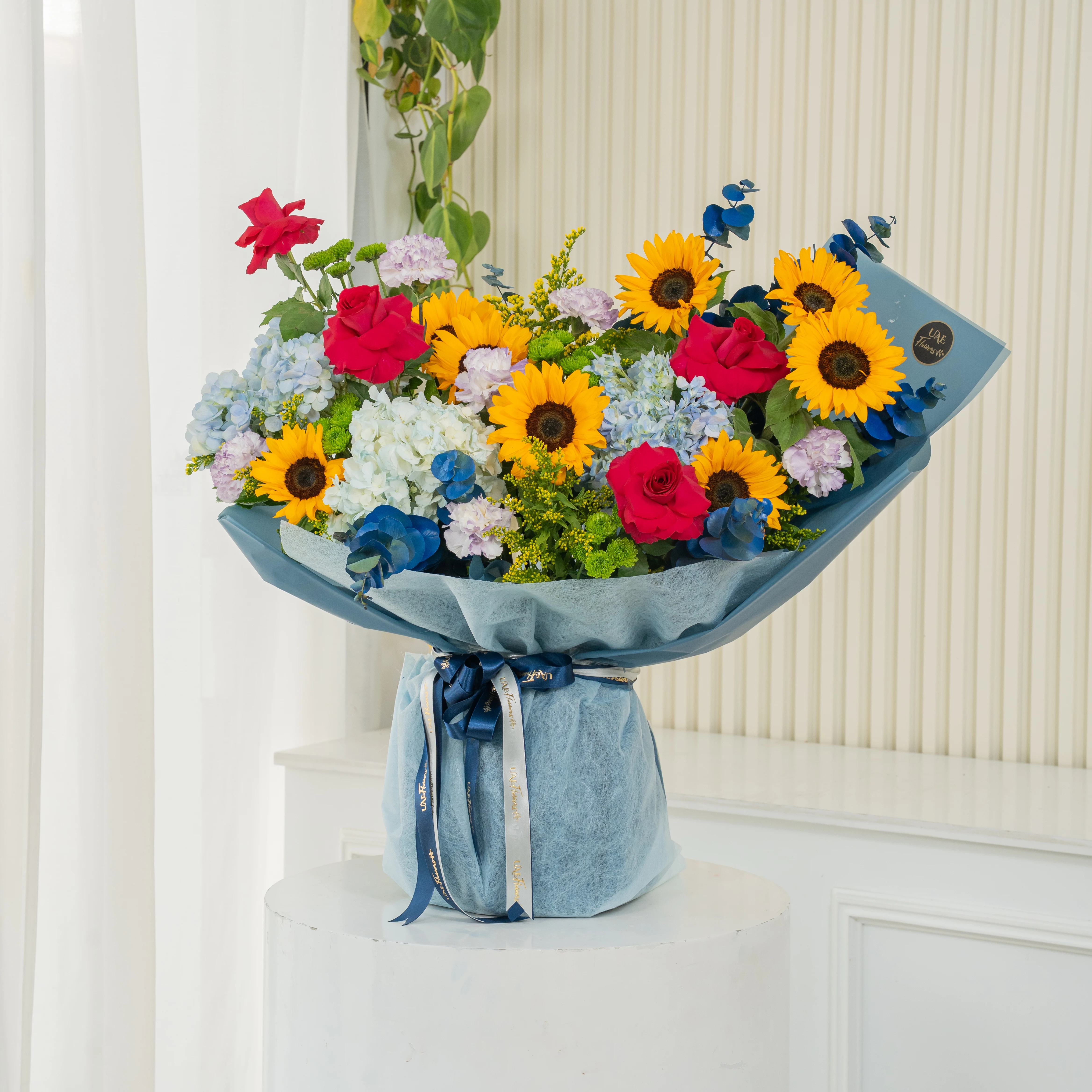 Bouquet of 8 sunflowers, 4 red roses, 5 blue hydrangeas, 6 carnations, green button chrysanthemums, blue sprayed eucalyptus leaf with green fillers in blue paper packing tied with blue ribbons.