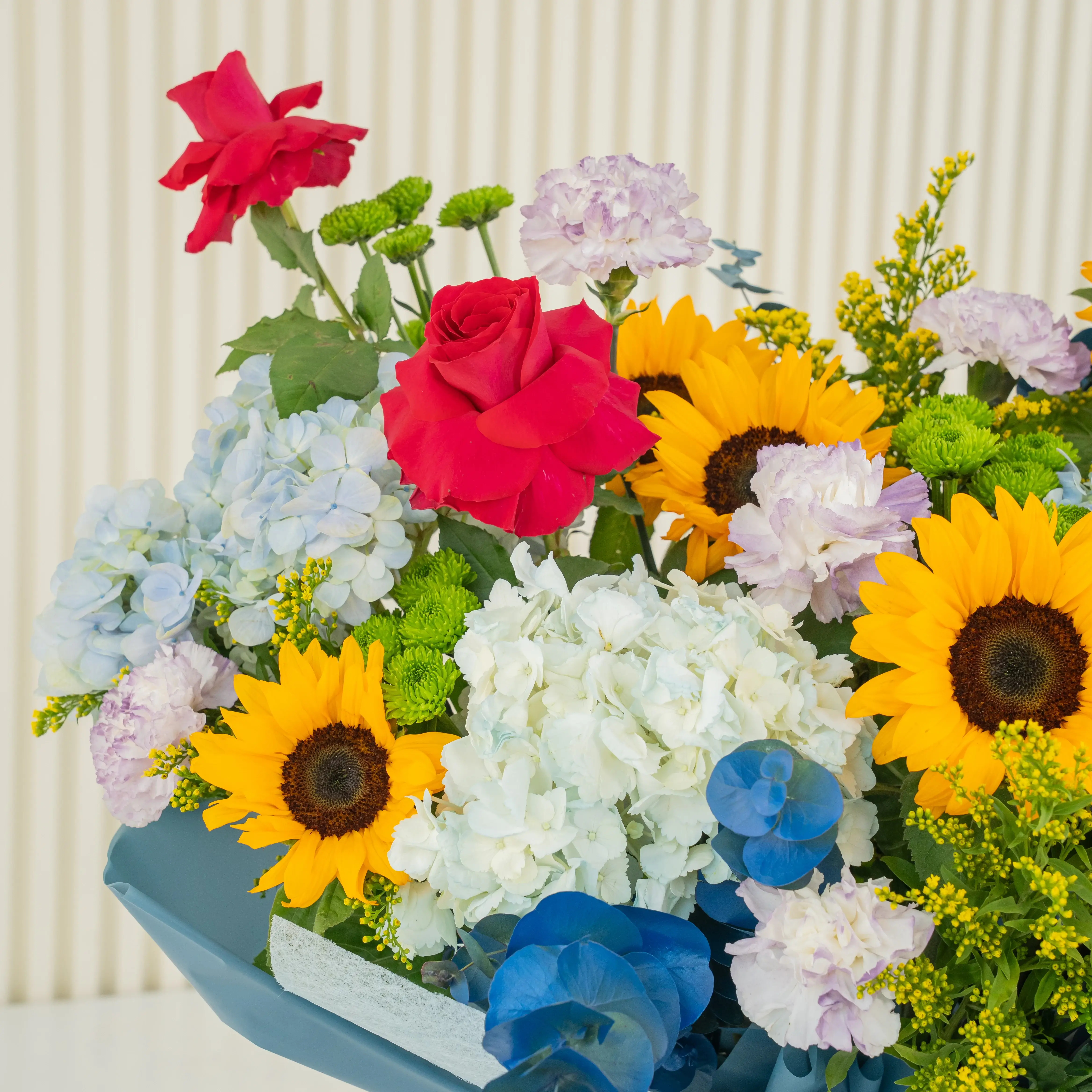 Bouquet of 8 sunflowers, 4 red roses, 5 blue hydrangeas, 6 carnations, green button chrysanthemums, blue sprayed eucalyptus leaf with green fillers in blue paper packing tied with blue ribbons.