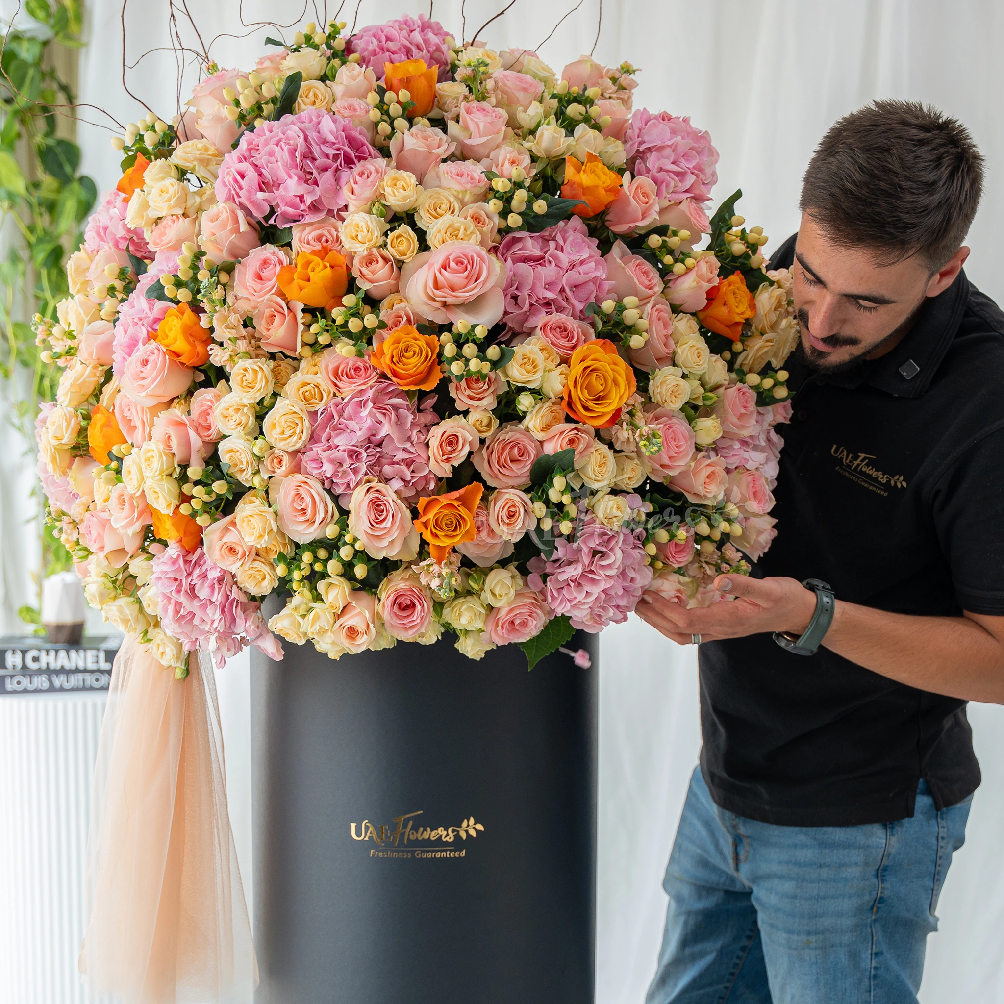 An arrangement of pink hydrangeas, pink roses, peach roses, orange roses, peach matthiola and white berries with green filler and peach net in a tall round box.