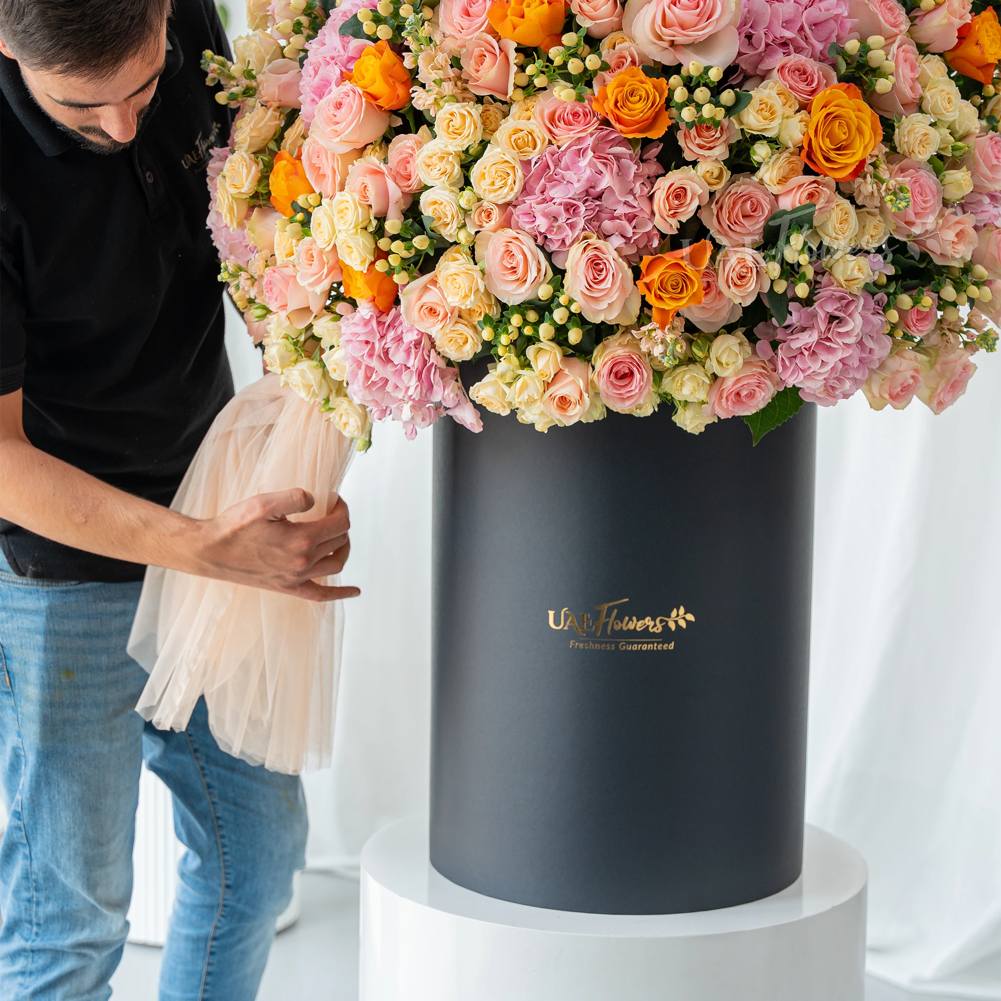 An arrangement of pink hydrangeas, pink roses, peach roses, orange roses, peach matthiola and white berries with green filler and peach net in a tall round box.