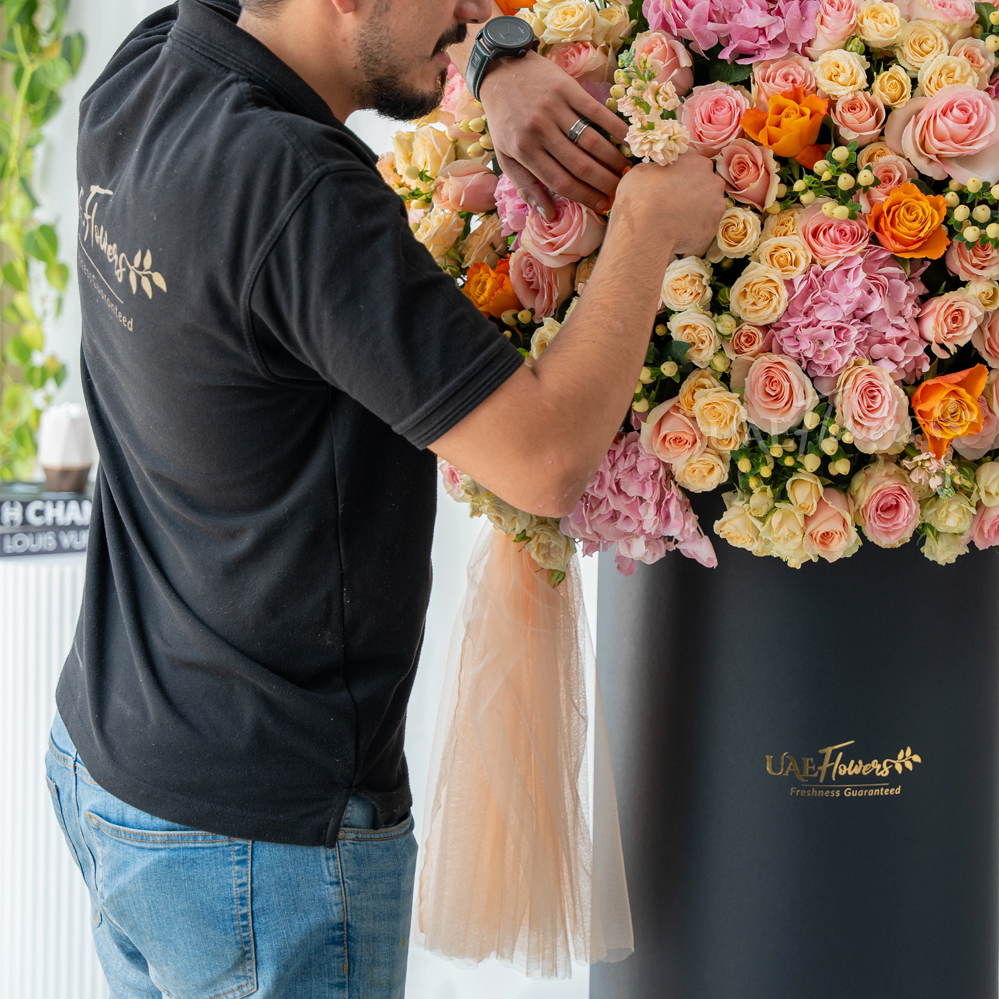 An arrangement of pink hydrangeas, pink roses, peach roses, orange roses, peach matthiola and white berries with green filler and peach net in a tall round box.