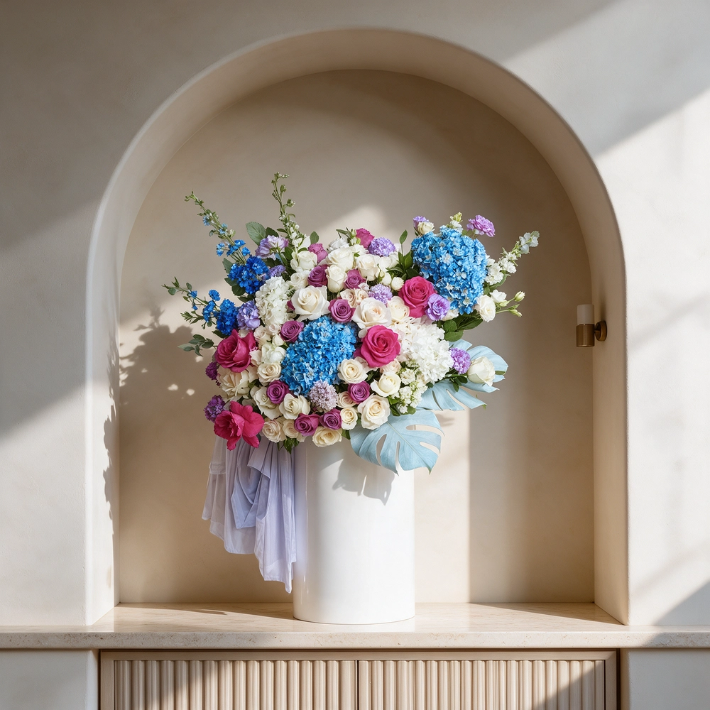 An arrangement of blue hydrangeas, white hydrangeas, purple roses, white roses, purple lisianthus, white spray roses, blue delphinium, fushia roses with green fillers and blue net in a tall round box.
