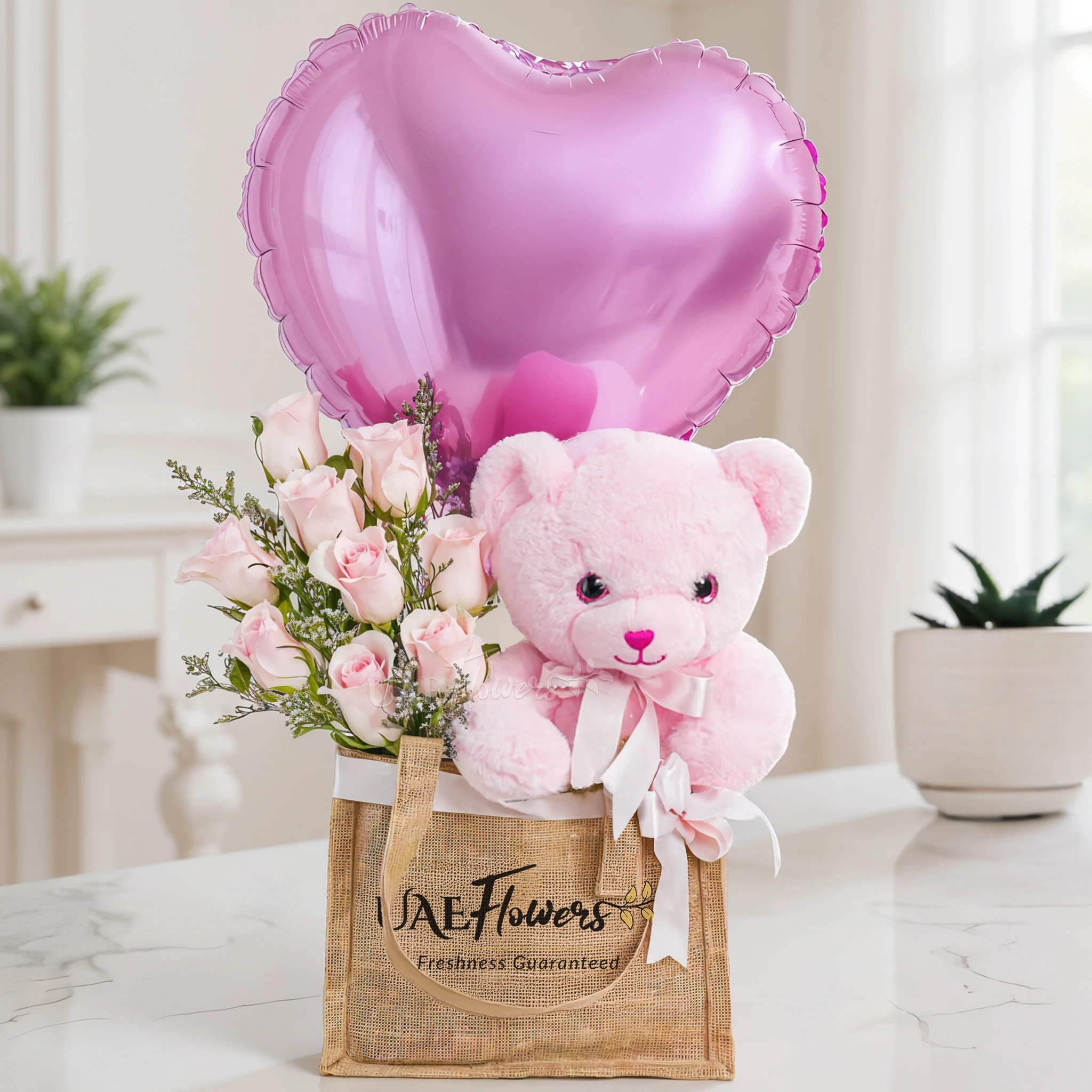 An arrangement of 7 pink roses, small size teddy bear and pink heart shaped balloons in a jute bag
