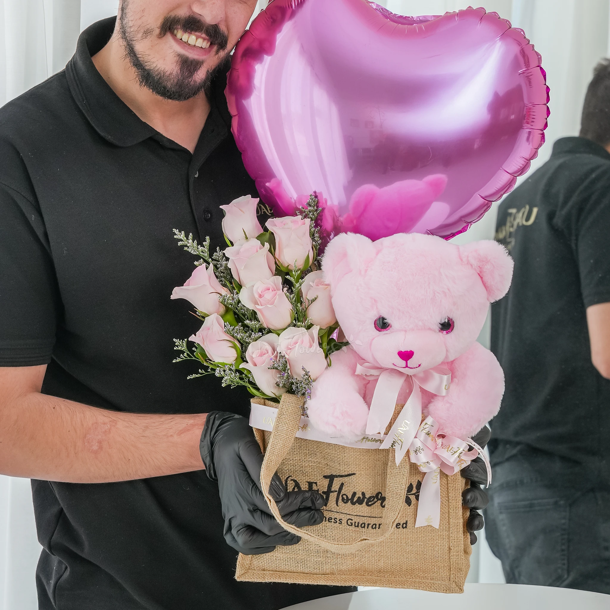 An arrangement of 7 pink roses, small size teddy bear and pink heart shaped balloons in a jute bag