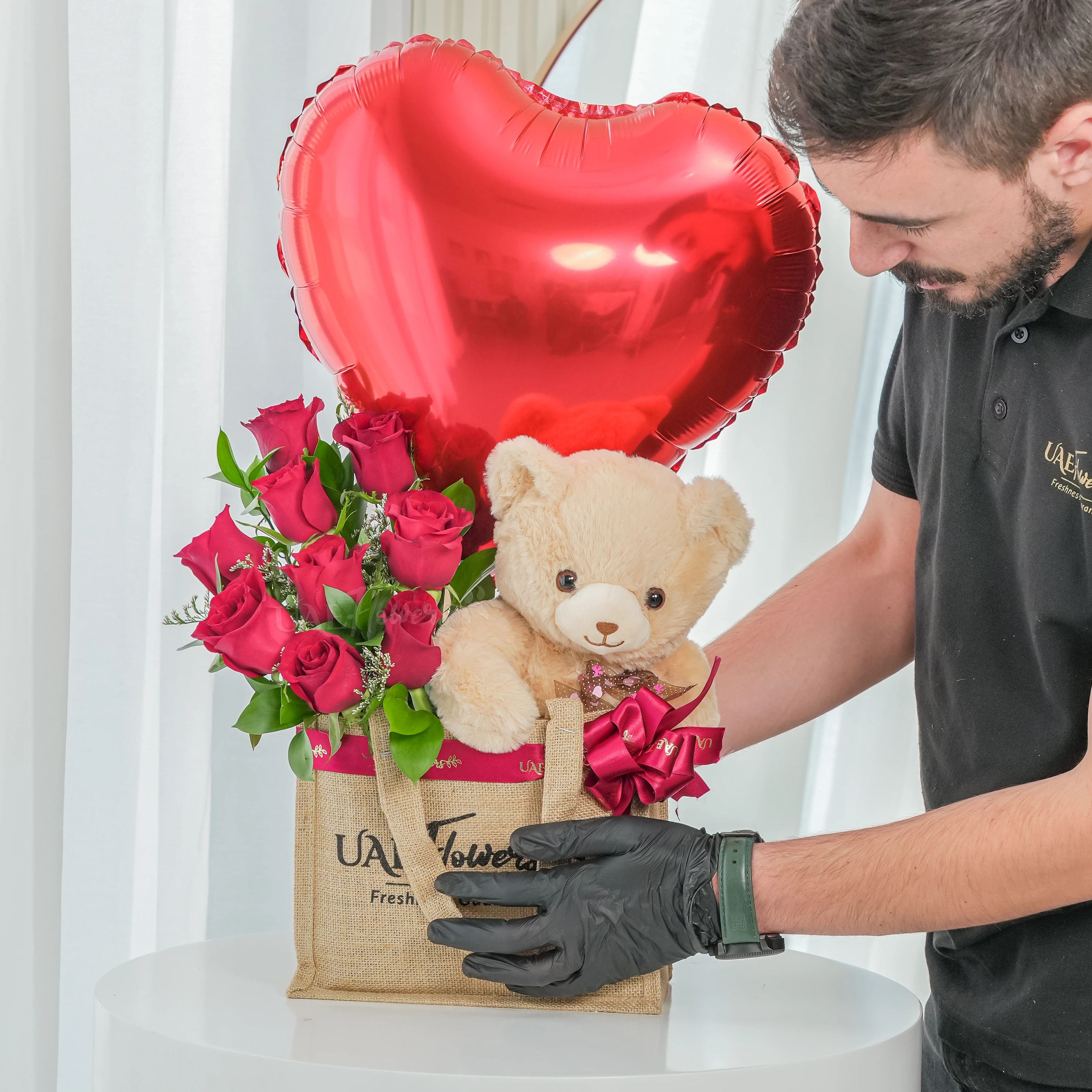 An arrangement of 7 red roses, small size teddy bear and hear shaped balloons in a jute bag