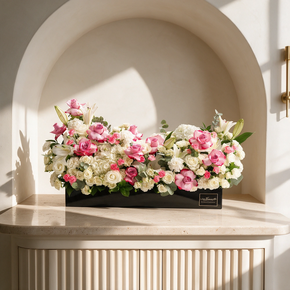 Arrangement of 5 white lilies, 15 purple roses, 15 white roses, 10 white carnations, 10 pink carnations, 10 white spray roses and 2 white hydrangeas with green fillers in a long black box.